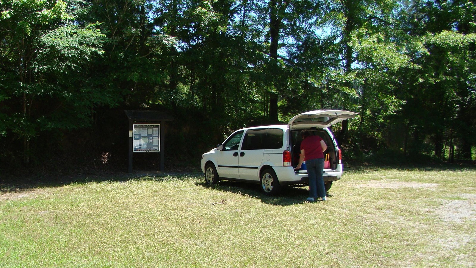 Trailhead parking in grass area. White minivan with the trunk open with a bulletin board behind.
