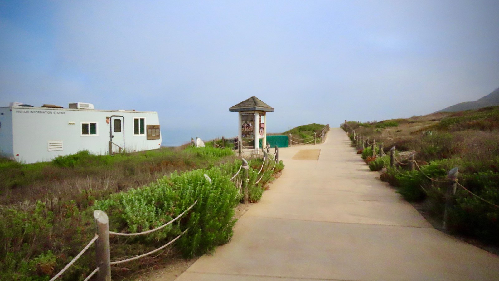 A tan concrete path with low lying bushes on each side. A kiosk and trailer are in the distance.