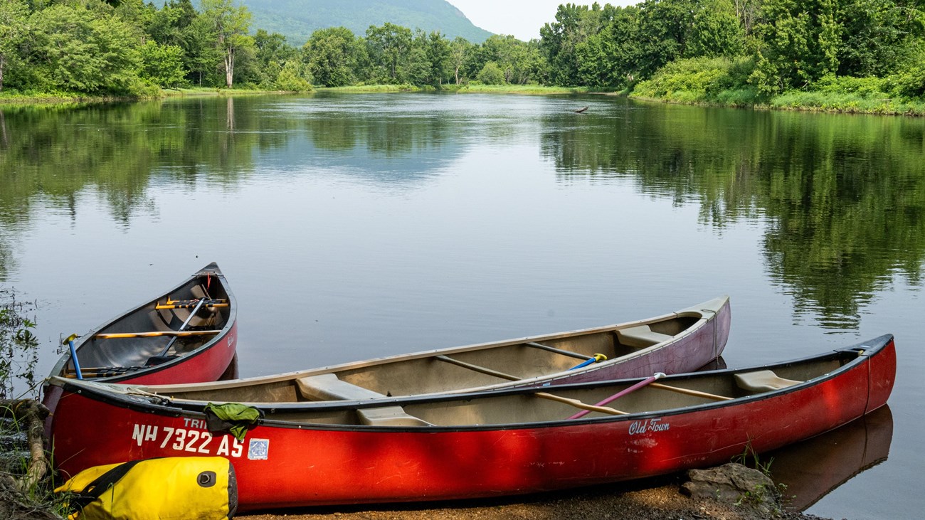 Three canoes sit at the dirt take-out of a campsite. The landscape reflects on the river.