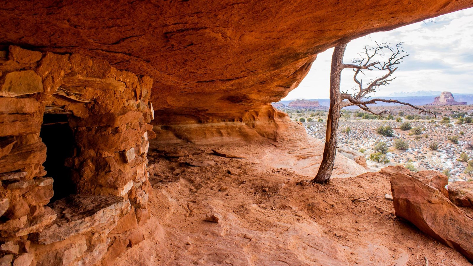 A stone and clay structure sits underneath an orange sandstone alcove on a cloudy day.