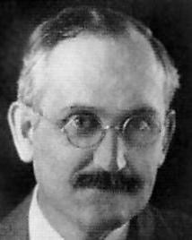 man with glasses and mustache wearing a suit poses for a picture. 