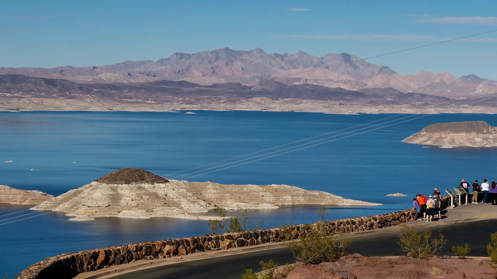 Scenic overlook parking lot with lake, islands, mountains in background. Cars and people. 