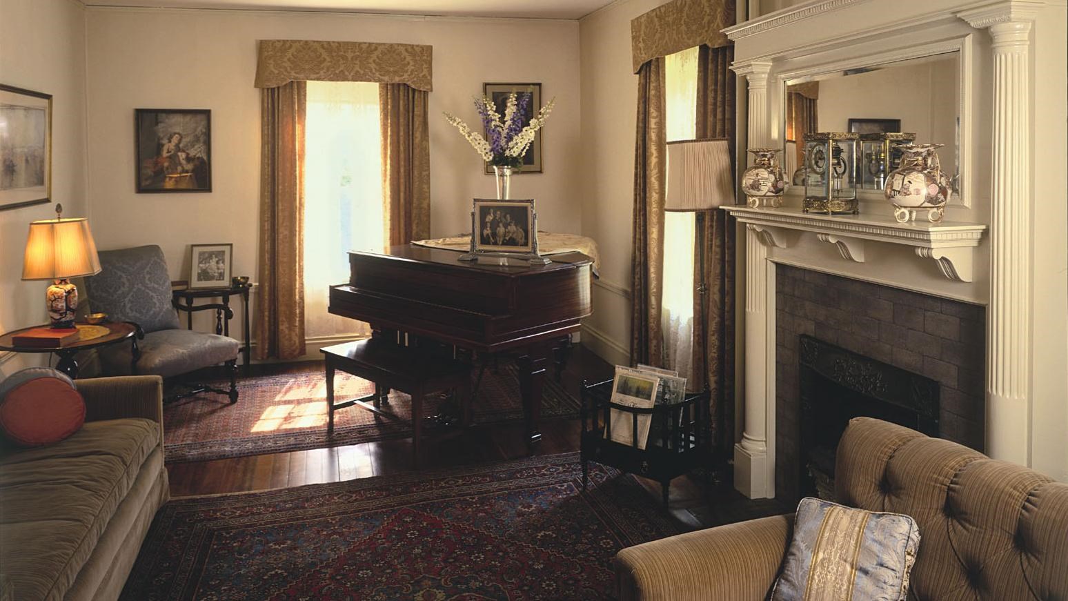 Long room with sofa and plush chairs arranged around tall mantle. Baby grand piano at back of room.