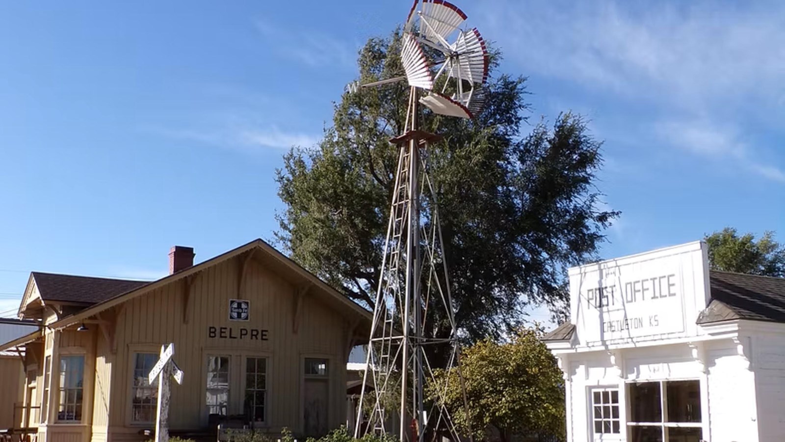 A historic wooden cabin sits next to a metal windmill and a small white, historic building.