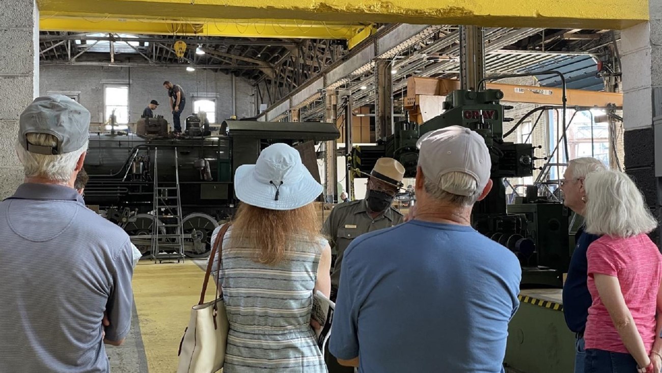 A ranger is shown speaking to a crowd of visitors while in a mechanical shop