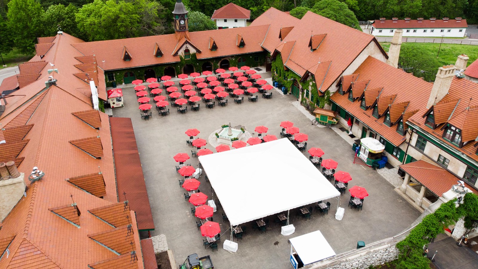 large courtyard with white pavilion, red umbrellas, and food & drink facilities with brown roofing. 