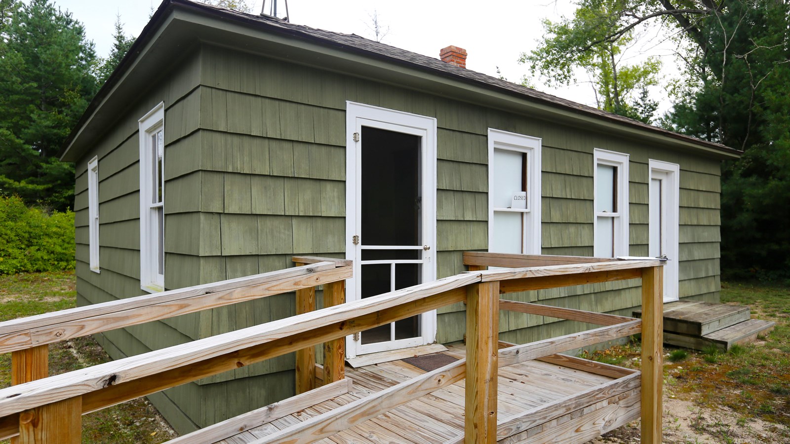 A one-story, green-shingled building with a sloping hip roof and double-hung windows