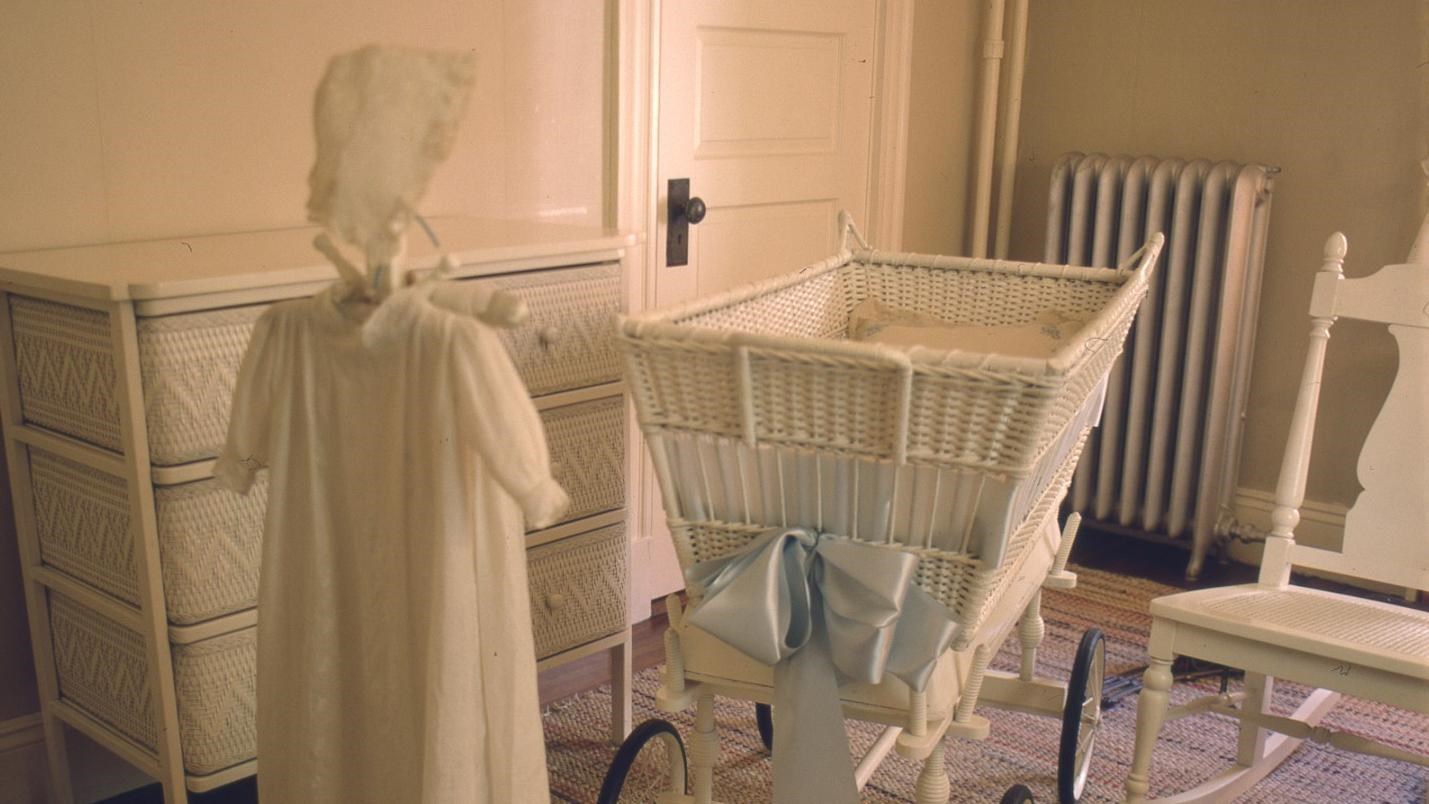 Room with white wicker furniture, including bassinet on wheels, and rag rug. Child\'s dress on stand.