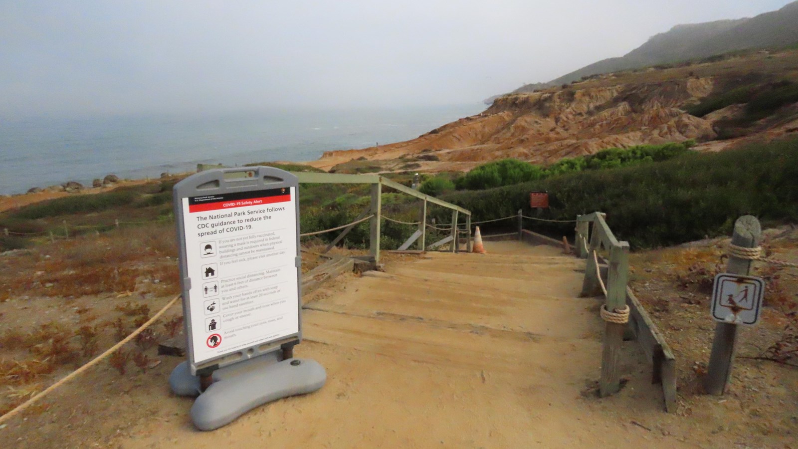 A dirt path overlooks the Kelp Forest in the ocean. A white sign is on the left of the trail.