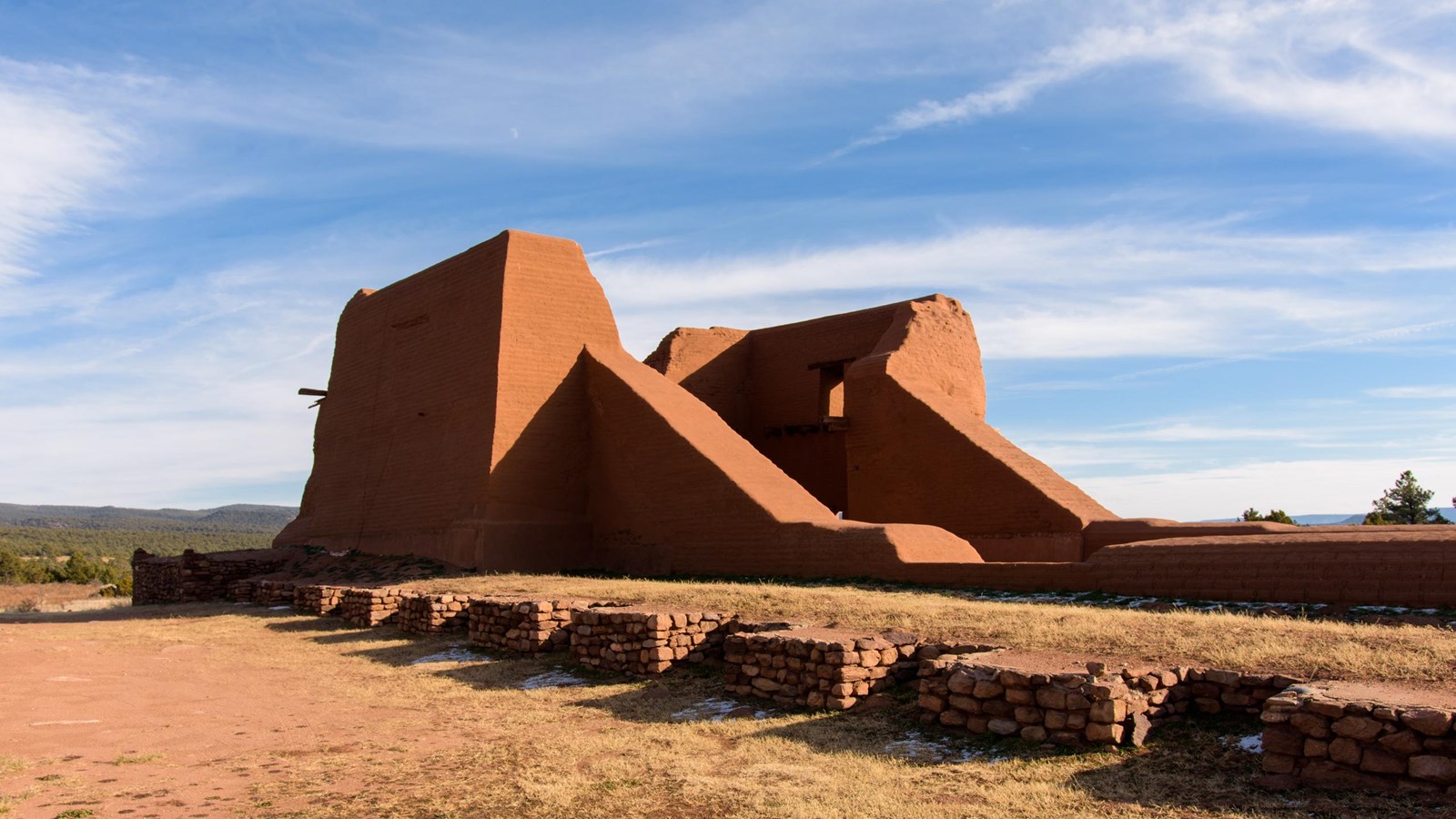 Large adobe ruins of a mission with blue skies, a few clouds, and trees in the background. 