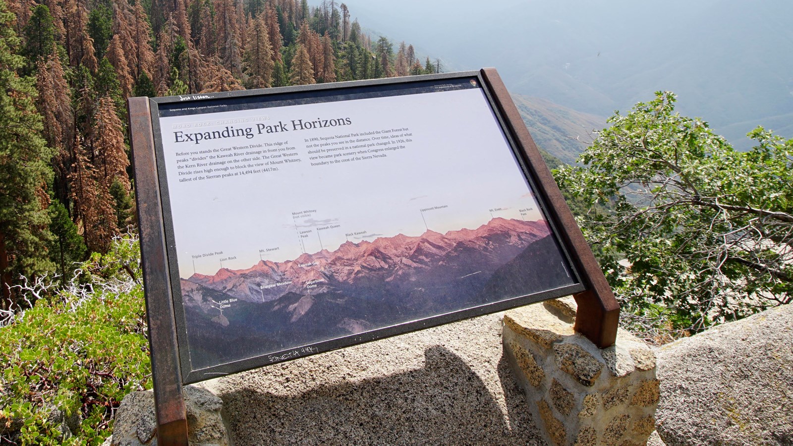 A metal panel sits in a metal frame. In the distance tall trees and mountains can be seen.