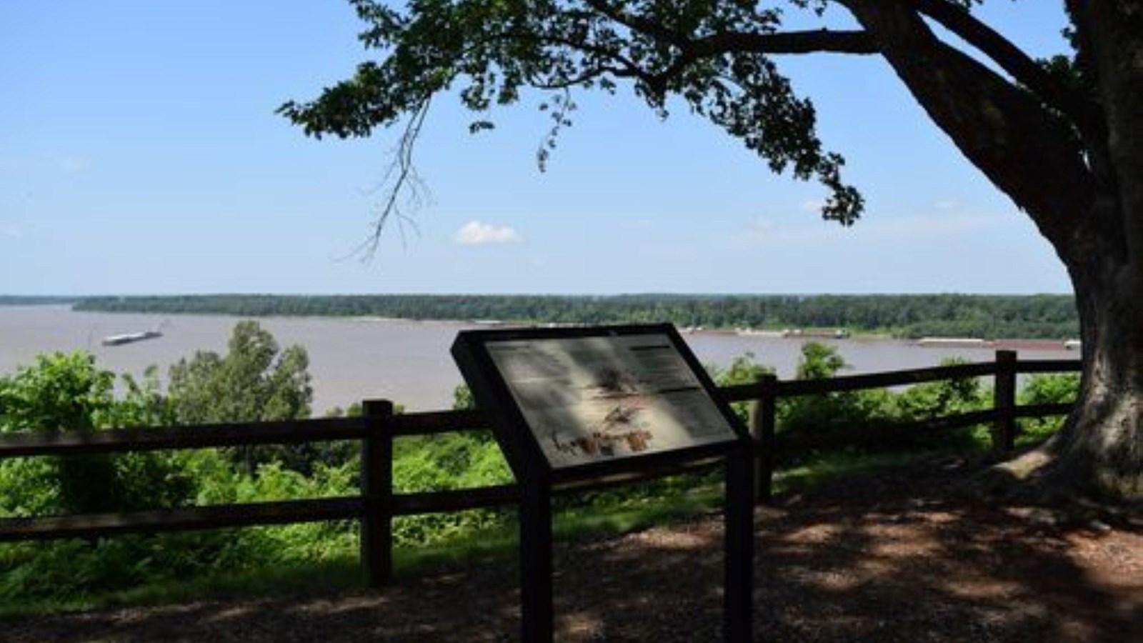 An exhibit sign sits next to a railing overlooking an expansive, flat river.