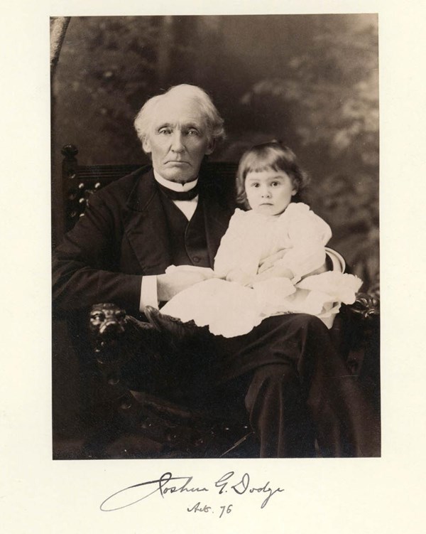 An older man wearing a black suit sits in a chair with a small child wearing white on his lap.