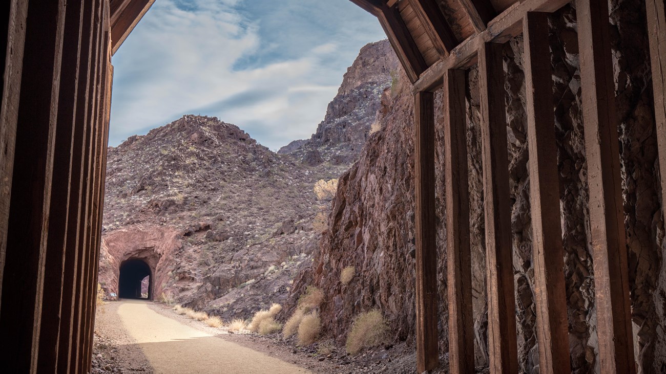 A tunnel constructed of wood beams looking out down a trail towards another tunnel.