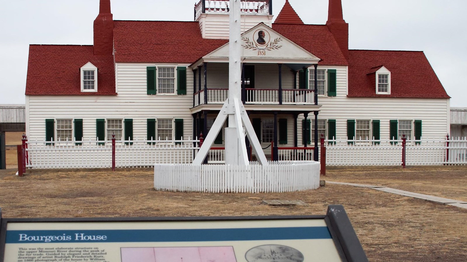 Exhibit with large white sided, red roofed house. White flagpole base between exhibit and house.