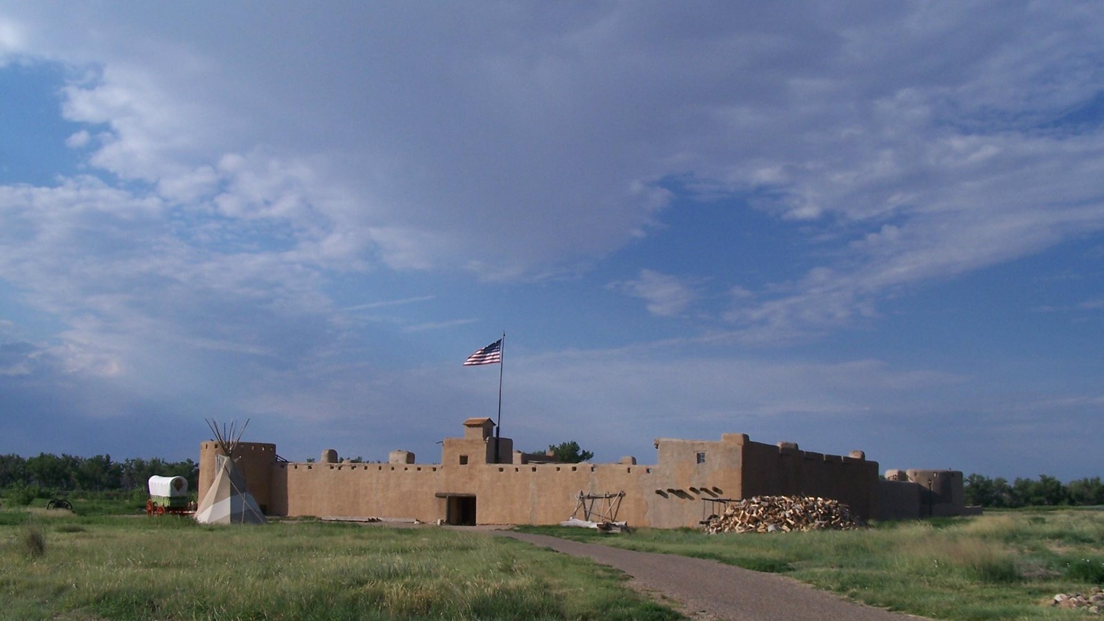 Large adobe fort with American flag flying above and a covered wagon, tepee, and path in front of it