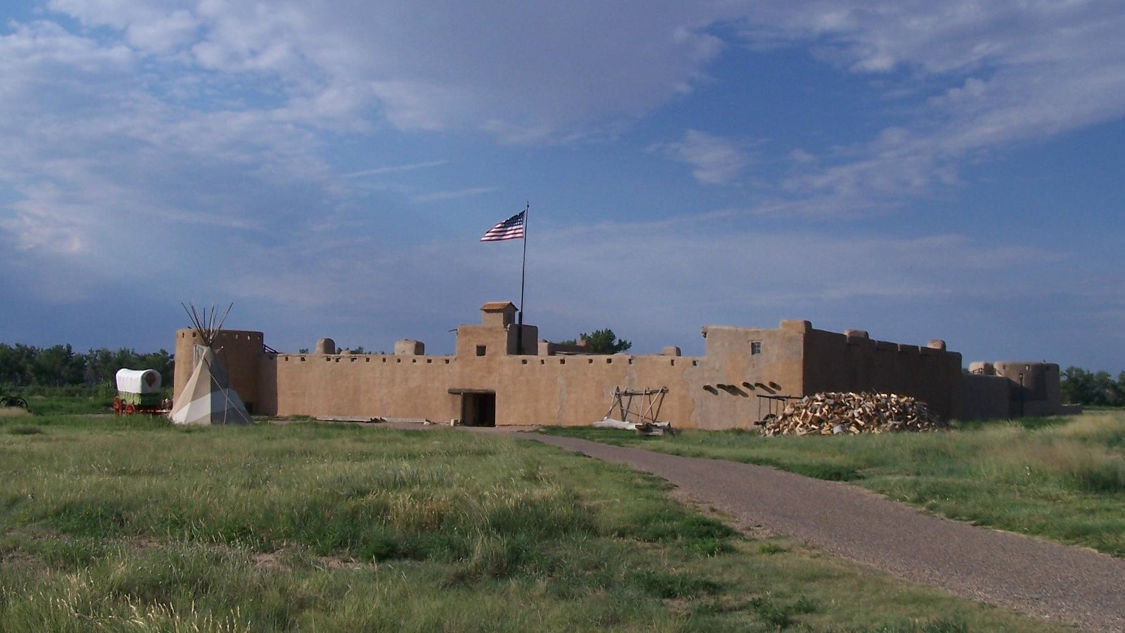 Large adobe fort with American flag flying above and a covered wagon, tepee, and path in front of it