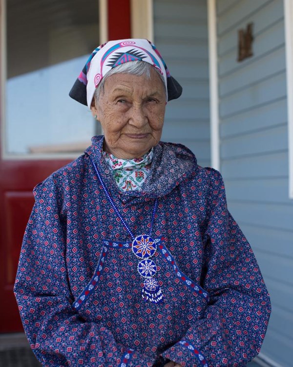 A woman sits on porch in full regalia with headscarf, kuspuk and rosette beaded necklace.