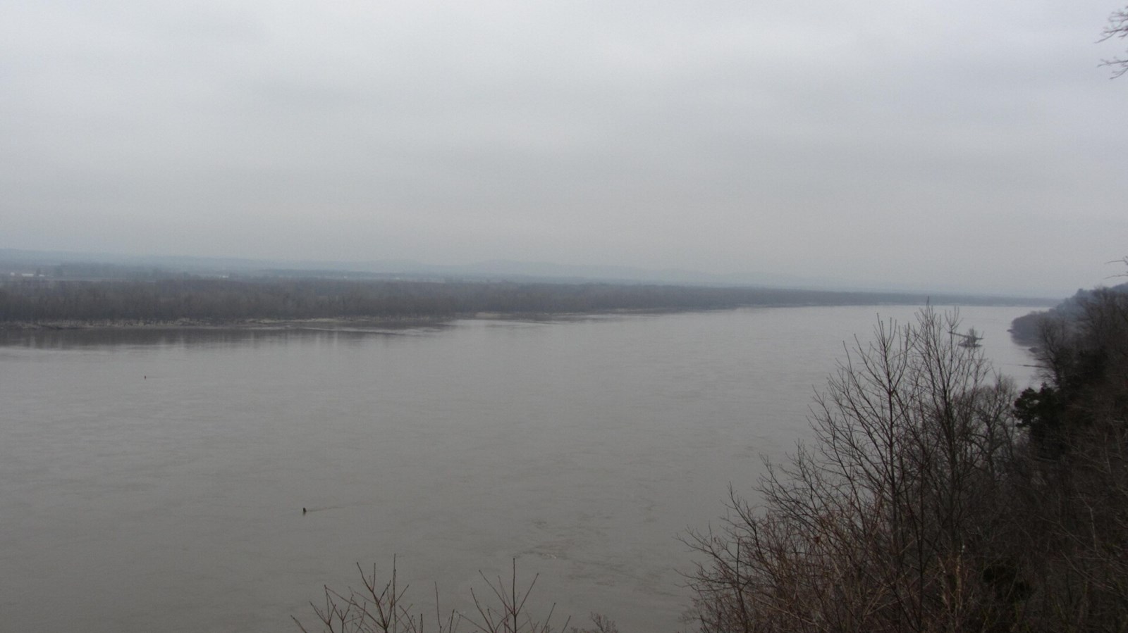 A hazy view of a large, flat river with vegetated banks.