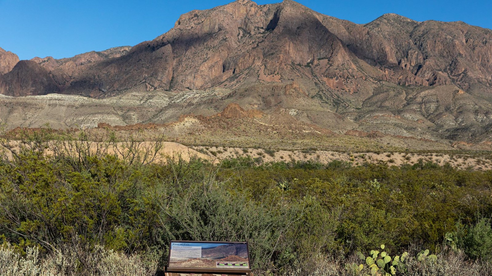 A metal sign sits on a rock base in front of a view of the Chisos Mountains