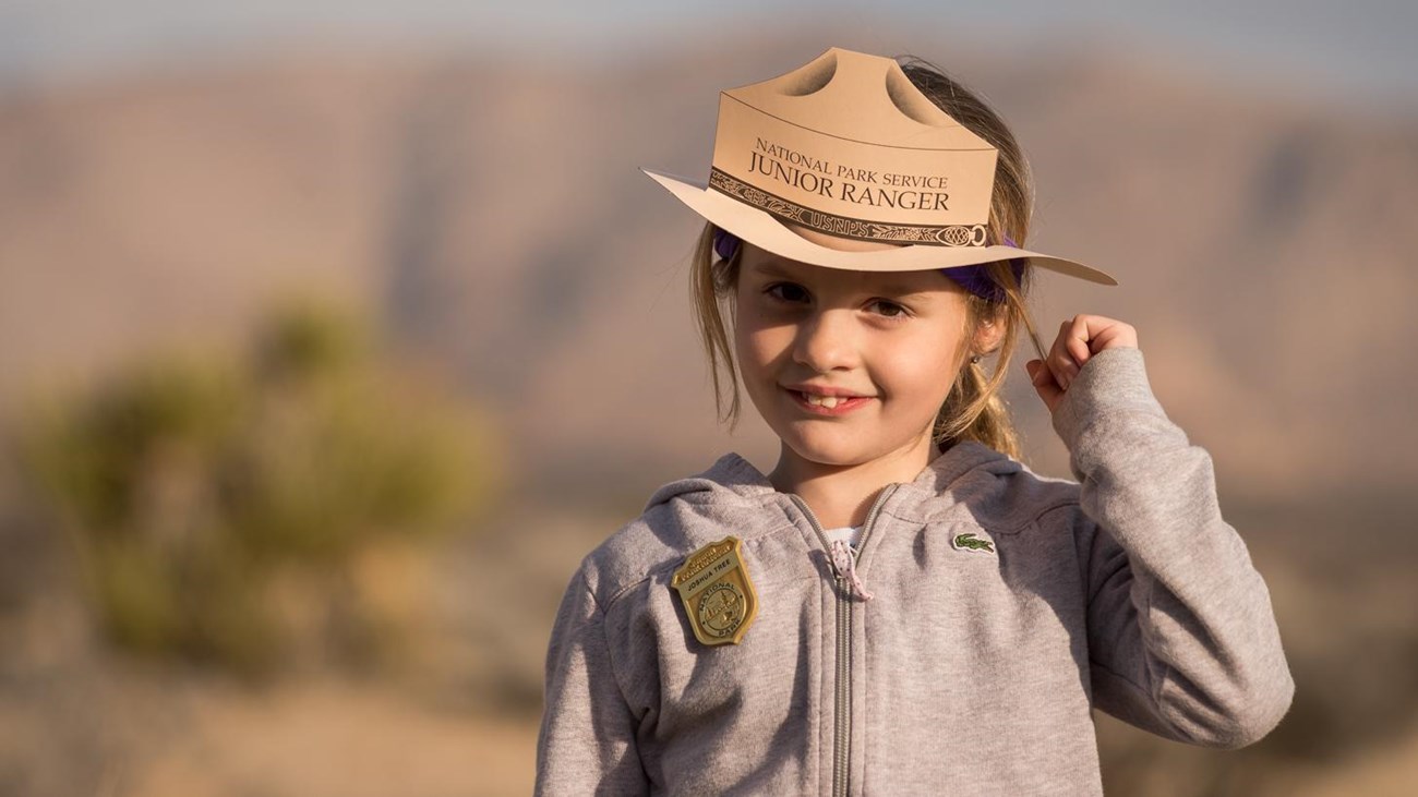 A young girl wearing a Junior Ranger badge and hat with mountains and a Joshua tree in the distance.
