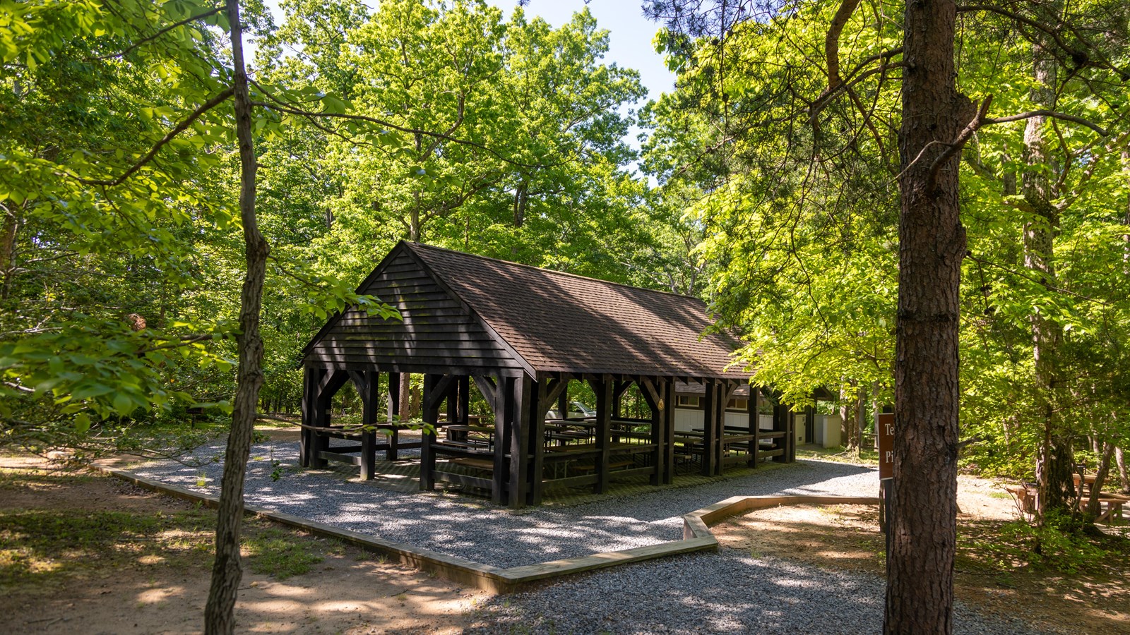 A wooden picnic pavilion stands among the woods. There are picnic tables under and surrounding it