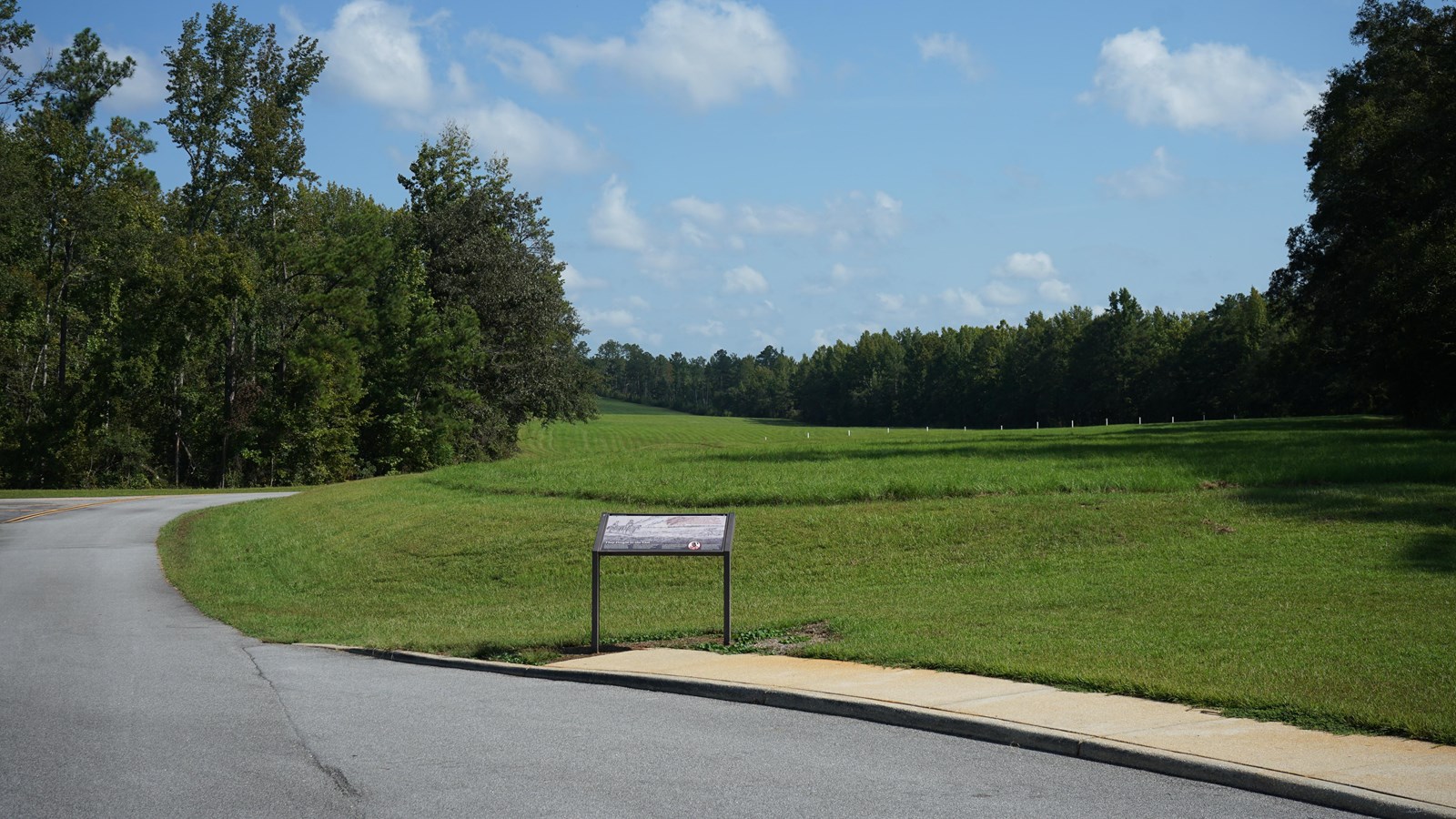 Image displays an informational wayside surrounded by grass and a forested area.
