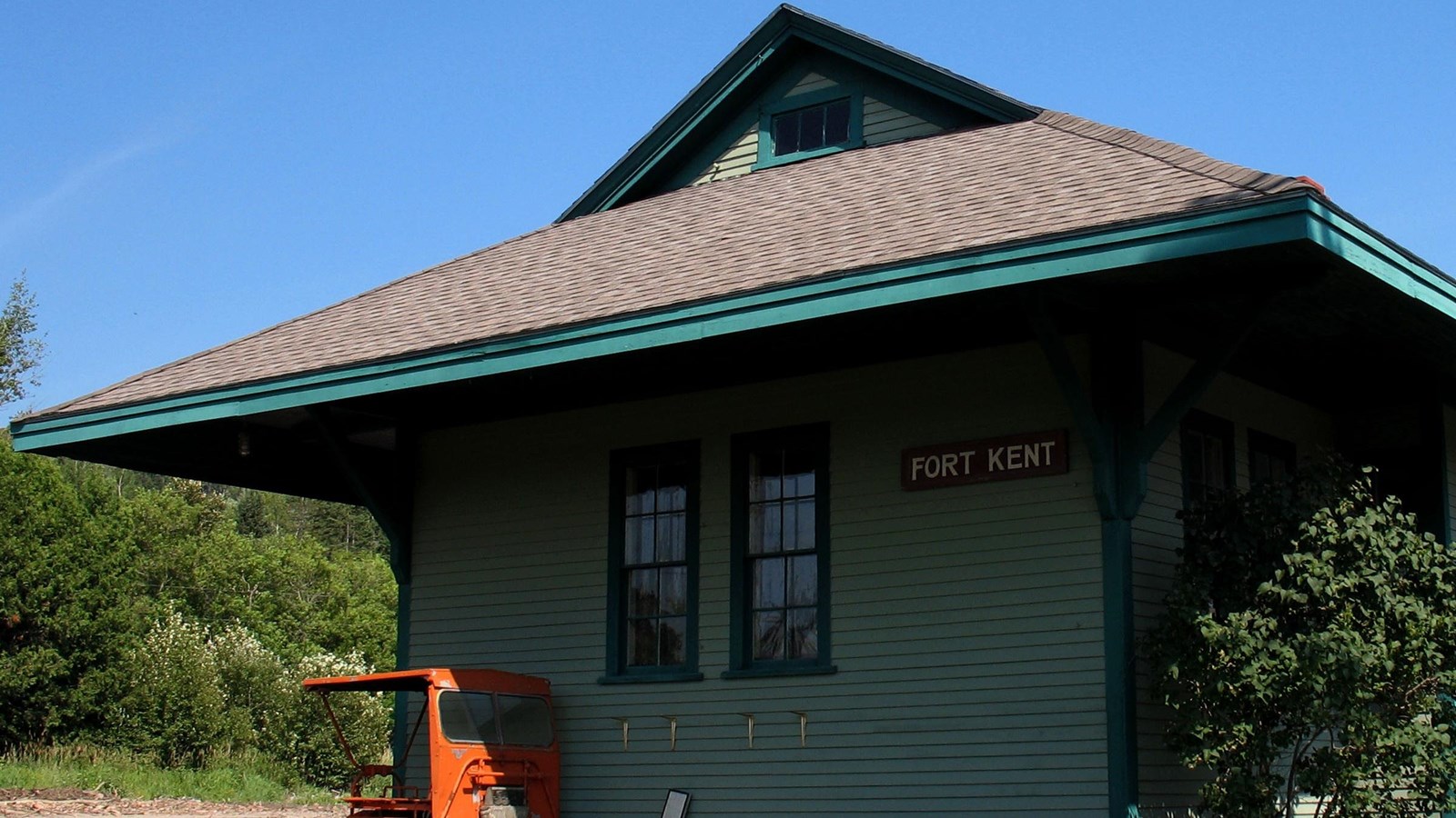 A small green wooden building with deep overhanging eaves. A small brown sign reads Fort Kent.