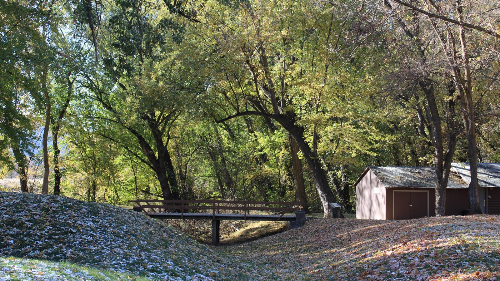Wooded scenic area with a wooden bridge crossing over a ditch filled with fall leaves.