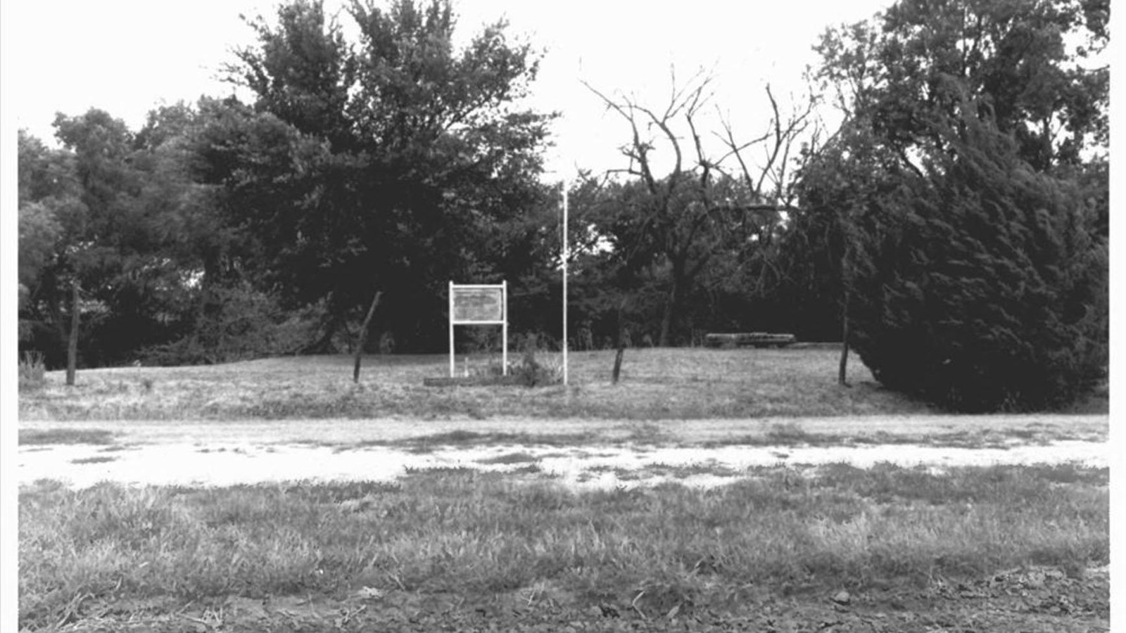 A historic black and white image of a grassy area with a sign and a flag pole.