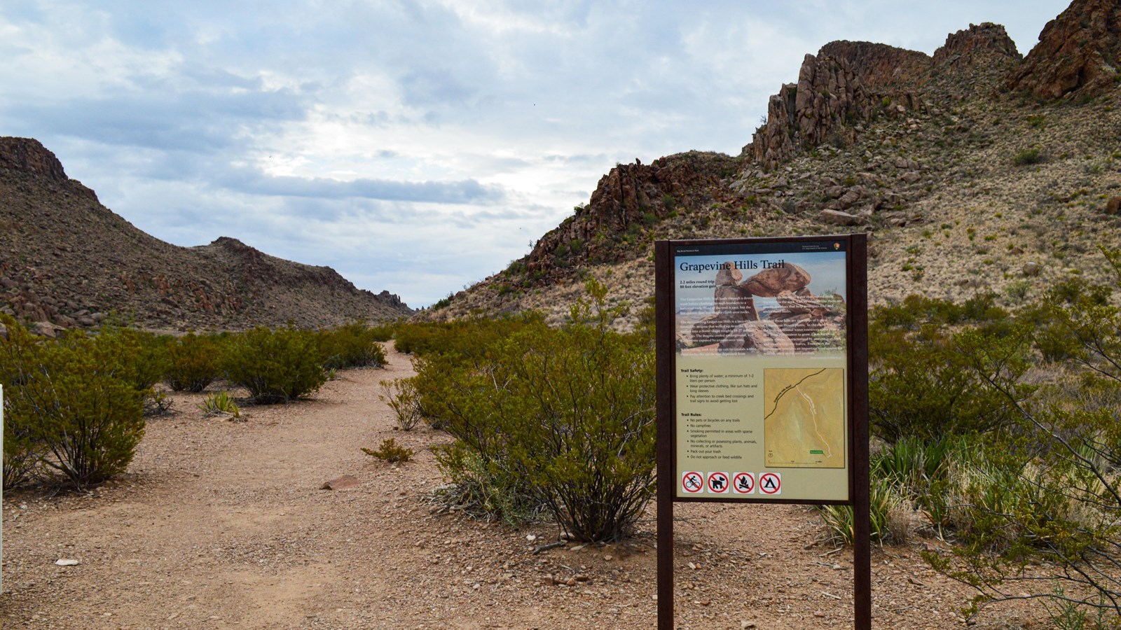 A metal sign with stands to the side of a gravel trail, with hills rising up around the trail.
