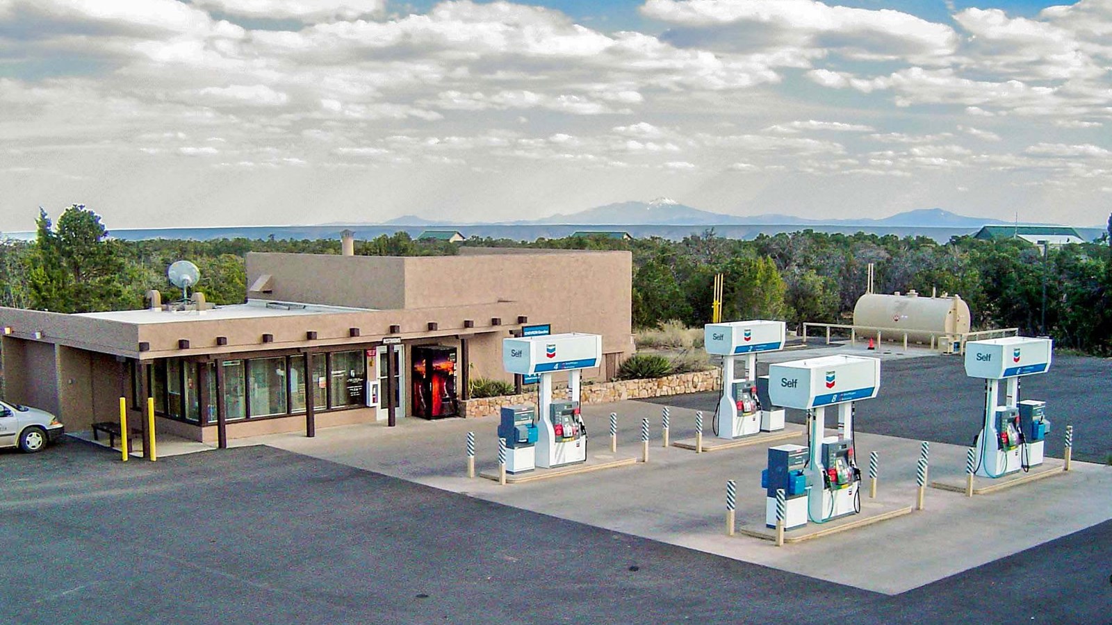 An adobe styled building sits on the edge of a large open area. Four gas pumps are out front.