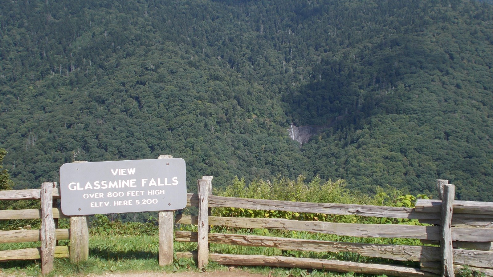 View of a mountain and a tall waterfall with overlook sign and wood fence in the foreground