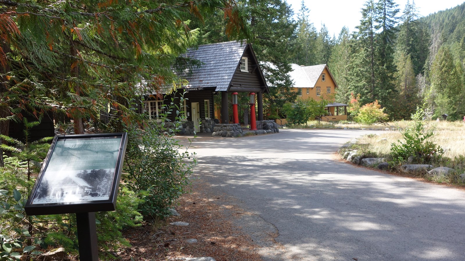A wayside panel next to a road overlooks a historic service station with red gas pumps.