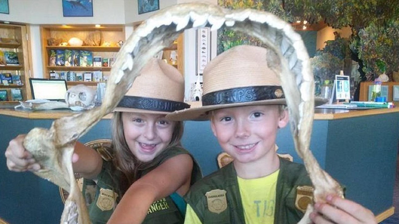 A girl and boy wearing ranger hats and badges peering out from a big shark jaw framing their faces