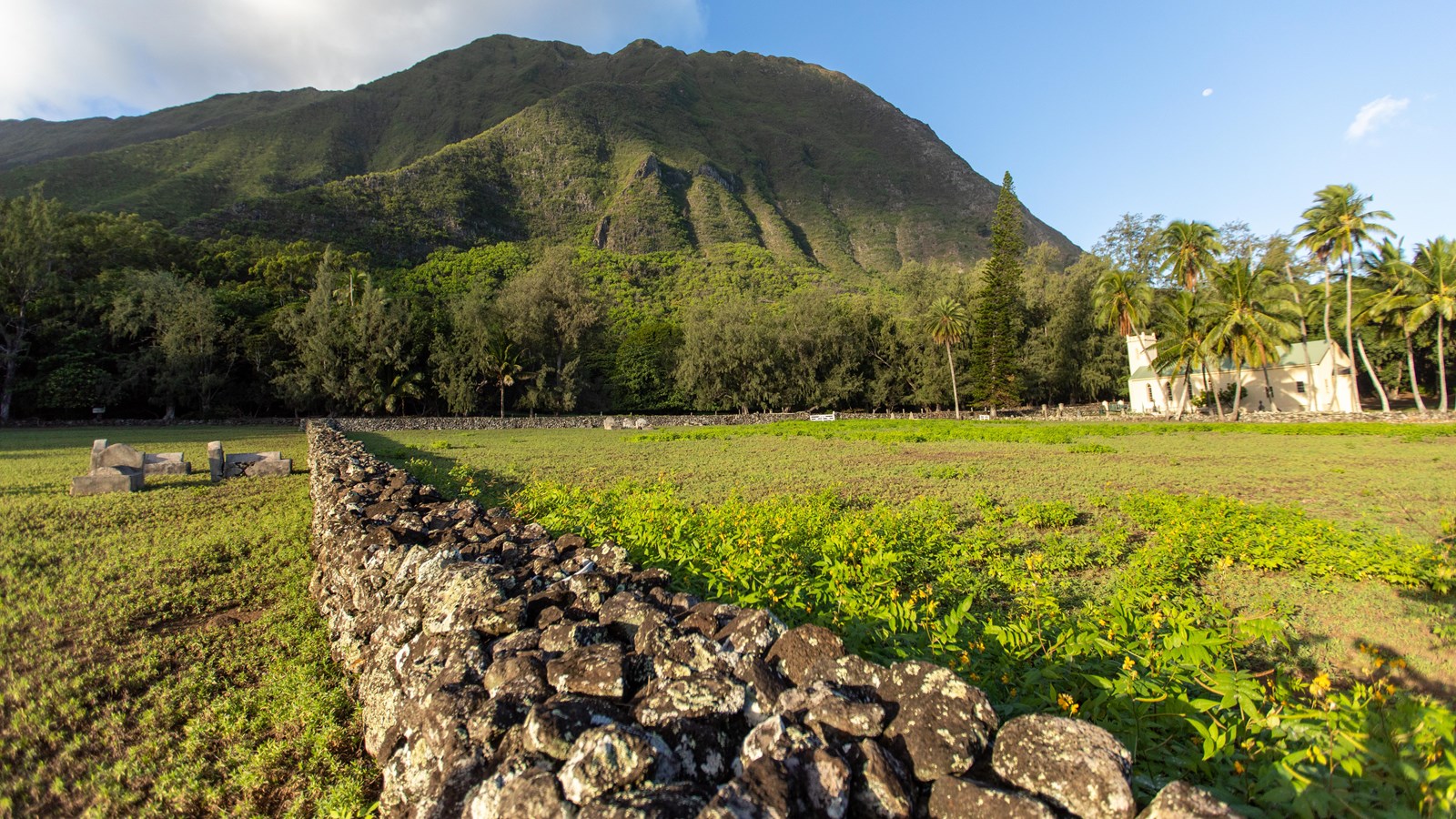A low rock wall surrounds a grassy field with pali in the background.