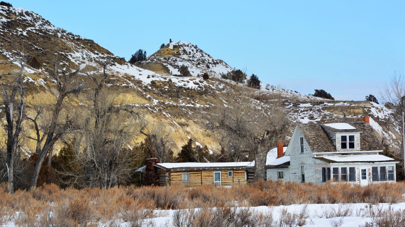 A winter scene, a white house sits next to a log building. Trees and buttes are behind them.