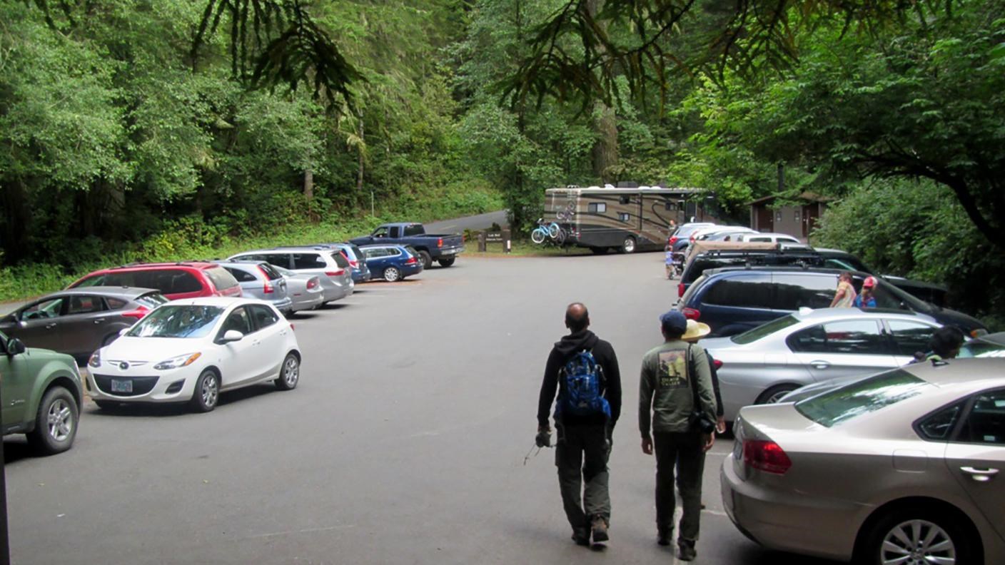 Vehicles parked in a small area. Two people walk away. Trees line the view.