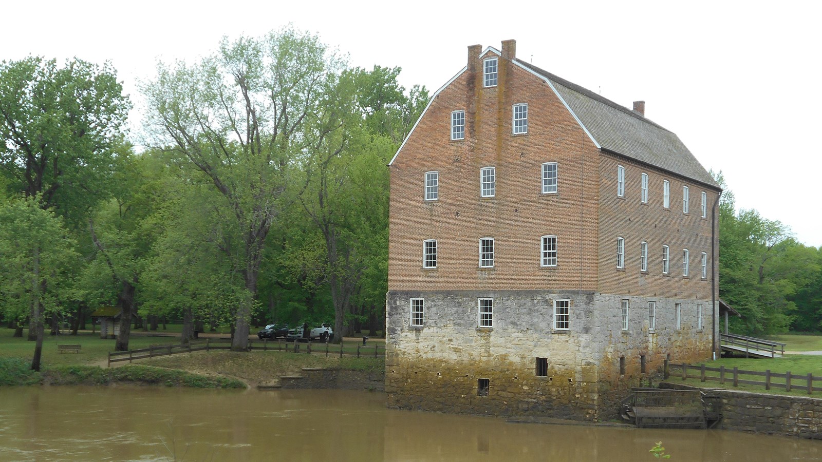 A large brick mill building, on the\ side of a muddy pond.