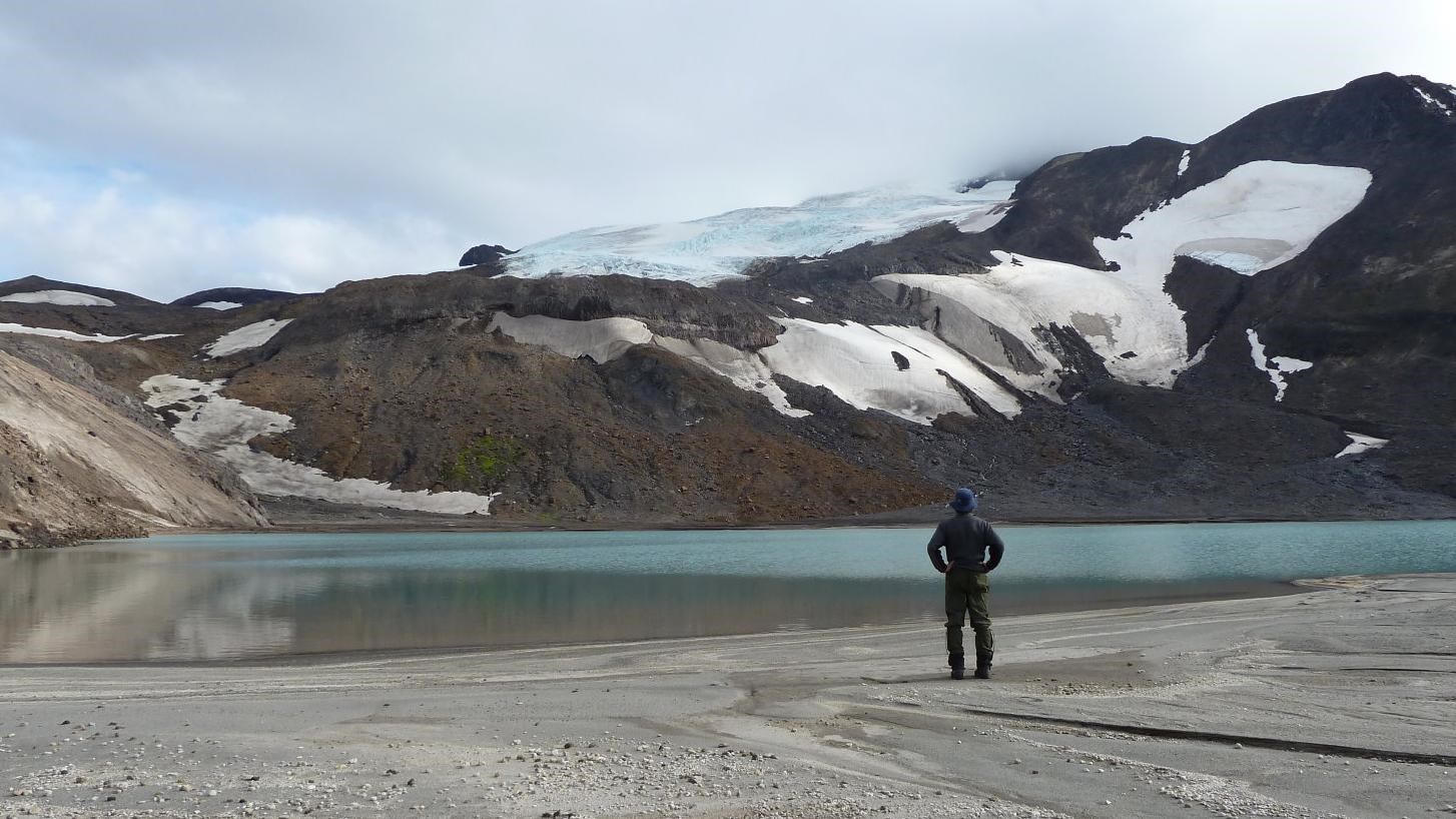 a person stands, hands on hips, beside a bright blue lake at the base of a snowy slope.
