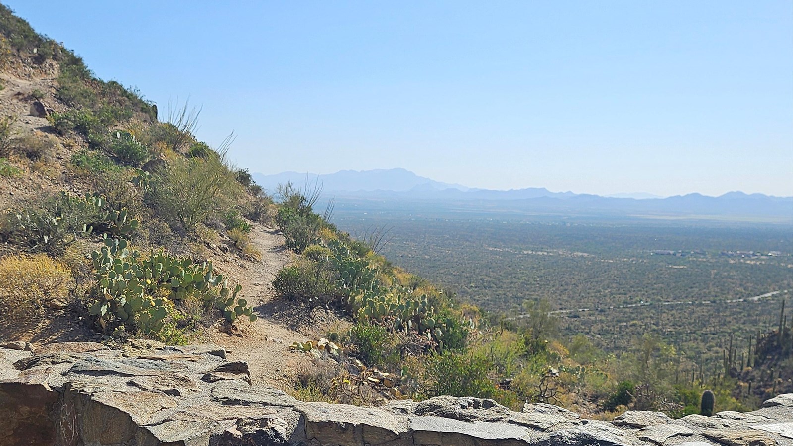 A rock wall lines the bottom of the image, a trail leads up a mountain to the left. A valley below