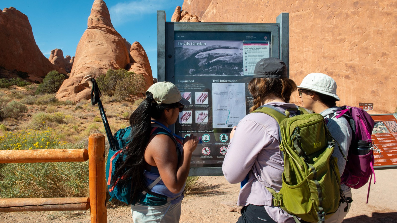 Hikers reading a trailhead sign near a fence, sandstone walls in background