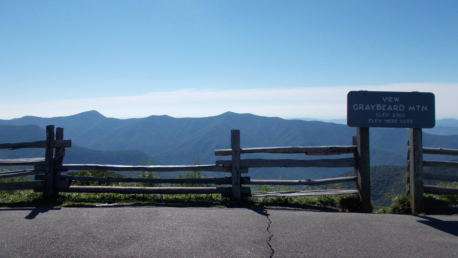 View of distant mountains with blue sky overhead and overlook sign, sidewalk and fence in foreground