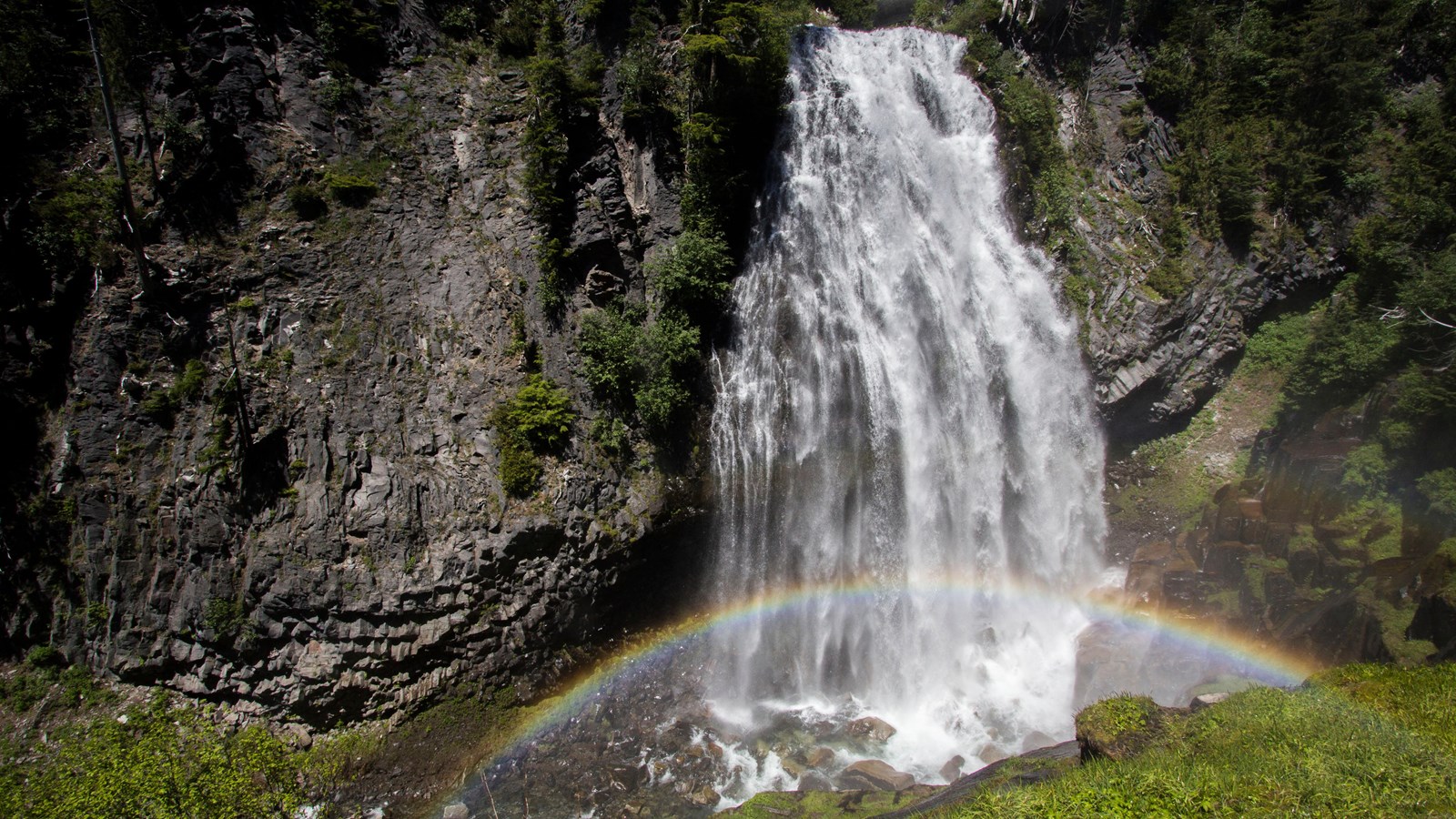 A tall waterfall gracefully plunges over a rocky cliff while a rainbow arches at its base.