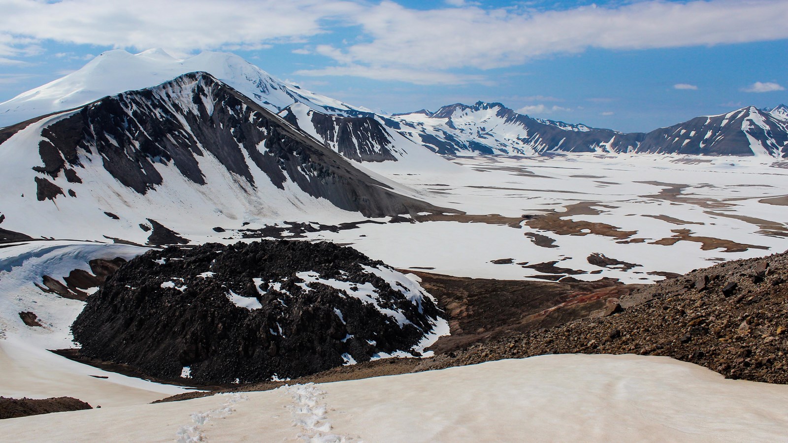A large, dark bump on the landscape sits amid a snowy mountain range.