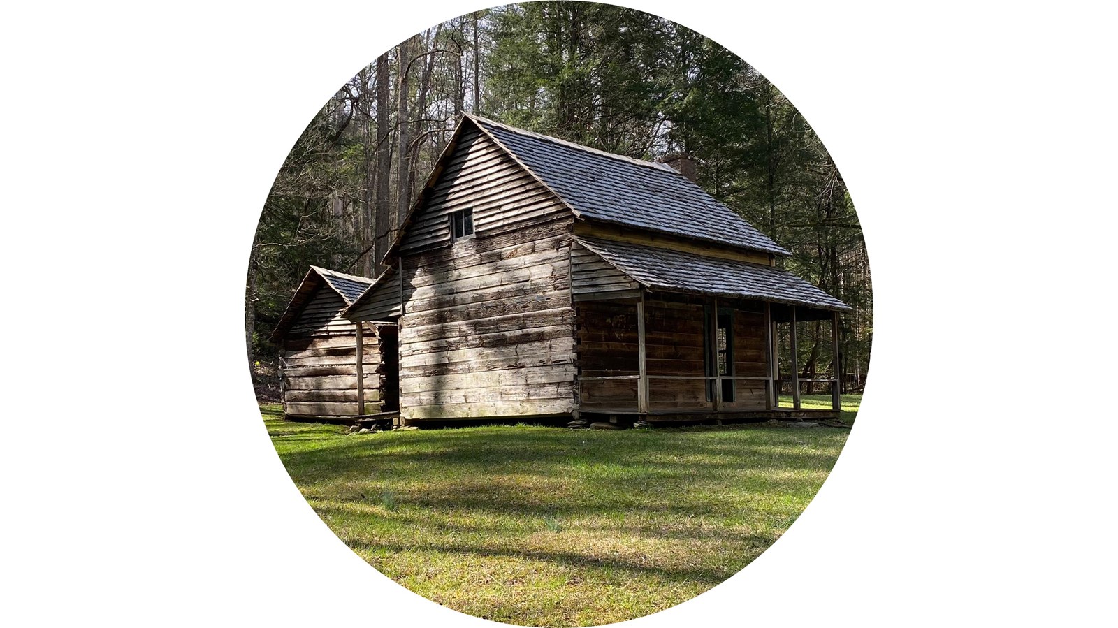 A log cabin and smokehouse on a grassy lawn surrounded by forest.