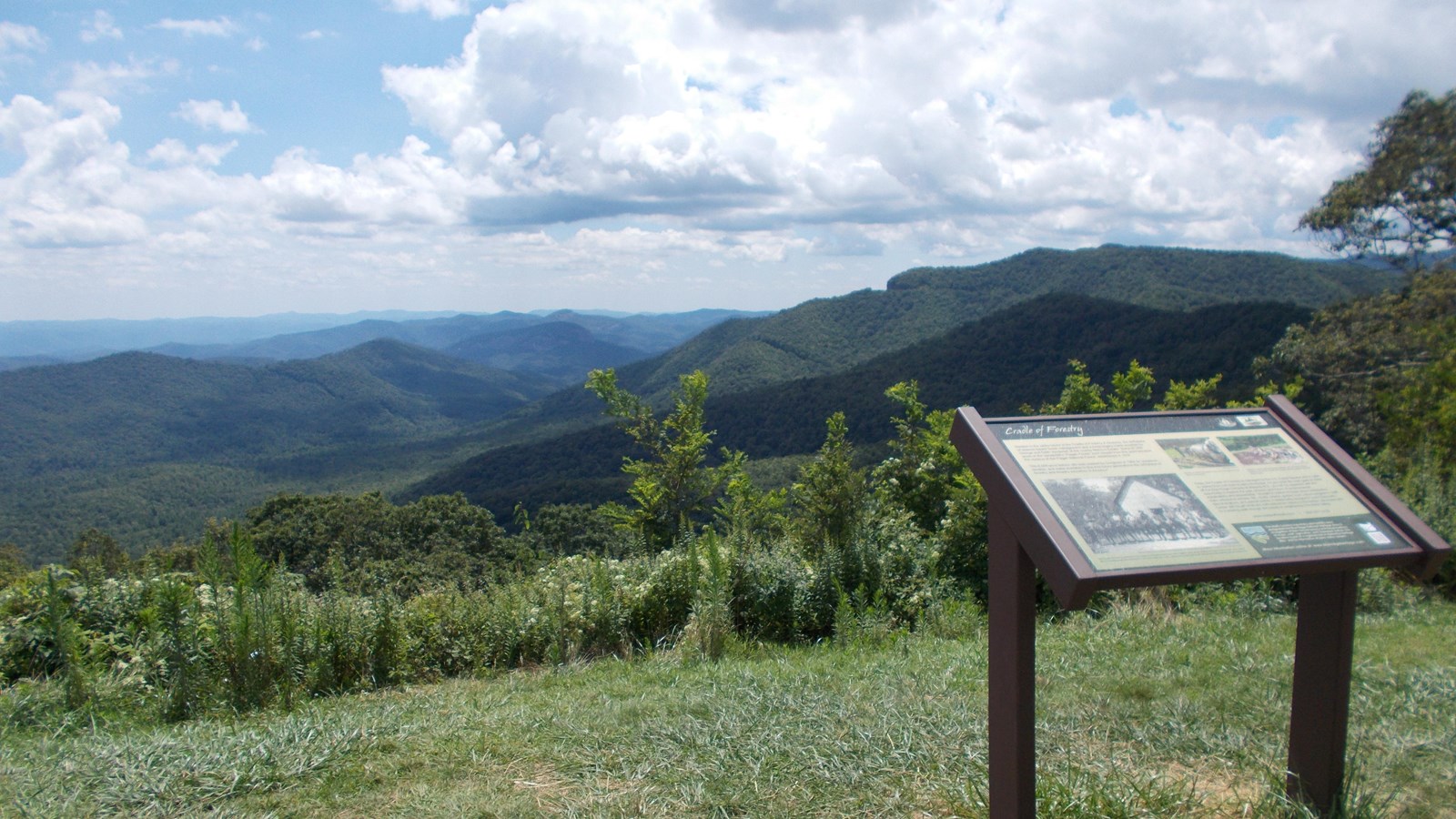 An information sign sits in front of an expansive view of green forested mountains 