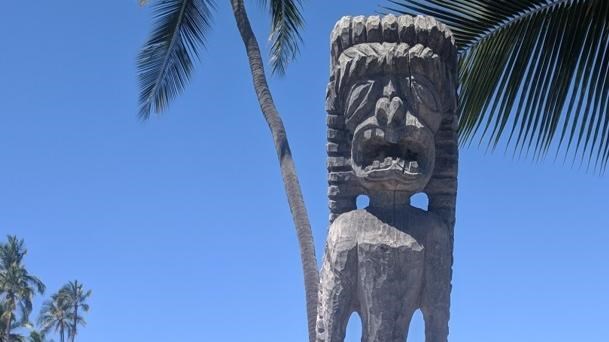 An intricately carved kiʻi (temple image) stands in the sandy royal grounds.