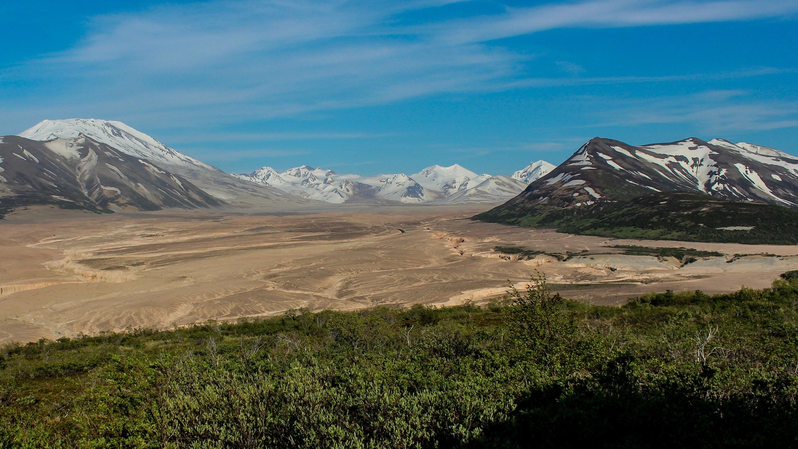 A large, tan, ash-filled valley is surrounded by snow covered mountains.
