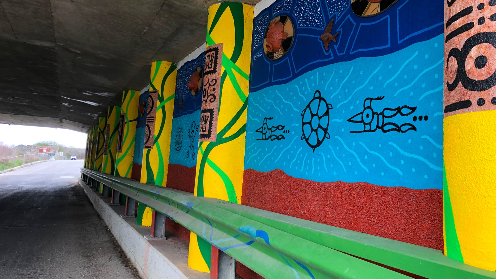 Yellow and green pillars in front of blue and red wall make up this underpass mural.